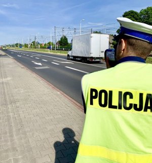 policjant stoi tyłem do fotografującego, trzyma w ręku miernik prędkości i mierzy prędkość samochodów poruszających się po drodze