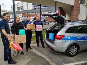 Policjanci trzymający pudełka z darami dla dzieci  stoją przy radiowozie z otwartym bagażnikiem