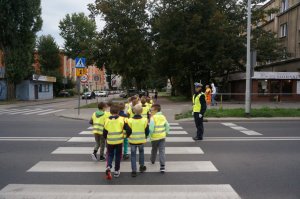 policjantka stoi przed przejściem dla pieszych, po którym przechodzi grupa dzieci w kamizelkach odblaskowych