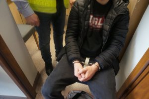mężczyzna siedzi na krześle ma założone kajdanki na ręce trzymane z przodu, przy nim stoi policjant ubrany w kamizelkę odblaskową