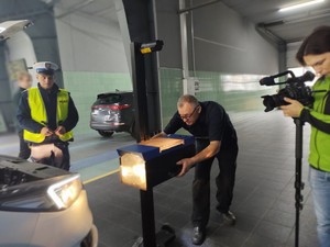 Kamerzysta i policjant przyglądają się jak diagnosta sprawdza oświetlenie samochodu